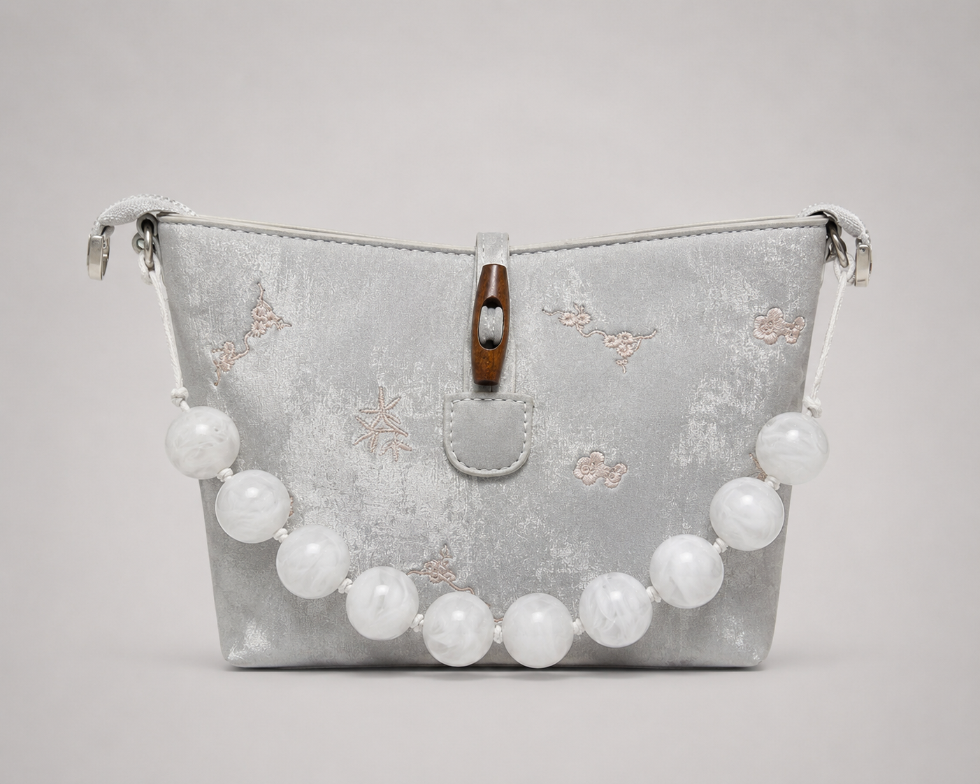 Front view of a silver metallic shimmer bag with a trapeze silhouette, featuring blush pink embroidered floral and star motifs, a chunky white marble resin bead carry handle, braided white rope shoulder straps with silver hardware, and a walnut wooden toggle closure at the top center.

